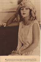 Norma Talmadge wearing white dress and pearls and a hat with white feathers across the front