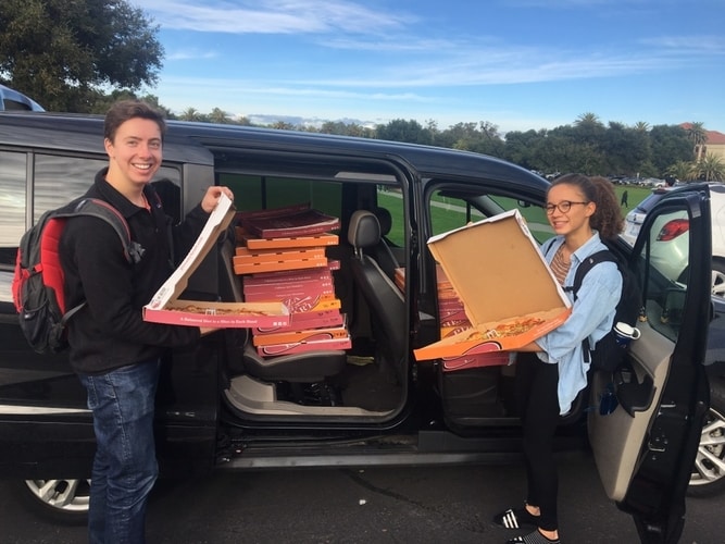 Two student volunteers standing by a van filled with pizza boxes
