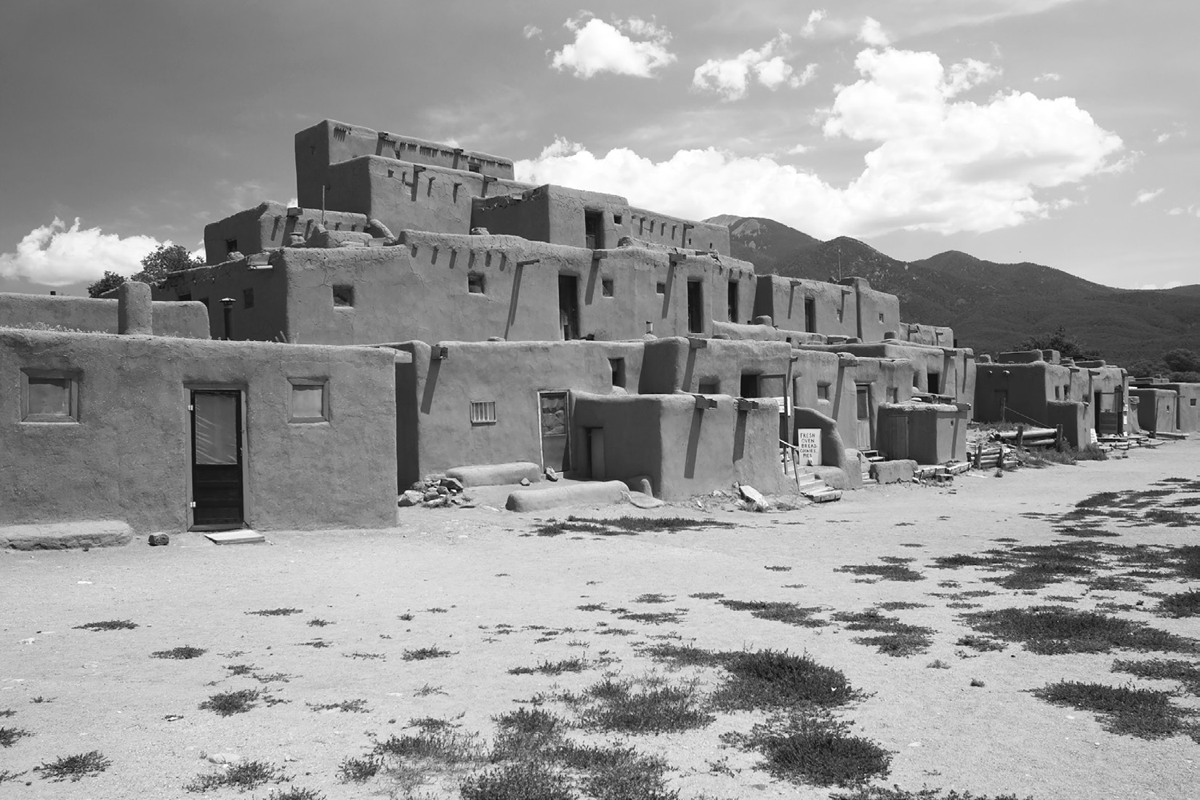 This 2007 photograph by Luca Galuzzi of Taos Pueblo shows a multistory adobe building complex in New Mexico. The ancient Native American settlement features characteristic tan-colored adobe with flat roofs, small windows, and wooden ladders connecting different levels. Bright turquoise door and window frames provide colorful accents against earth-toned walls. The foreground shows desert ground.