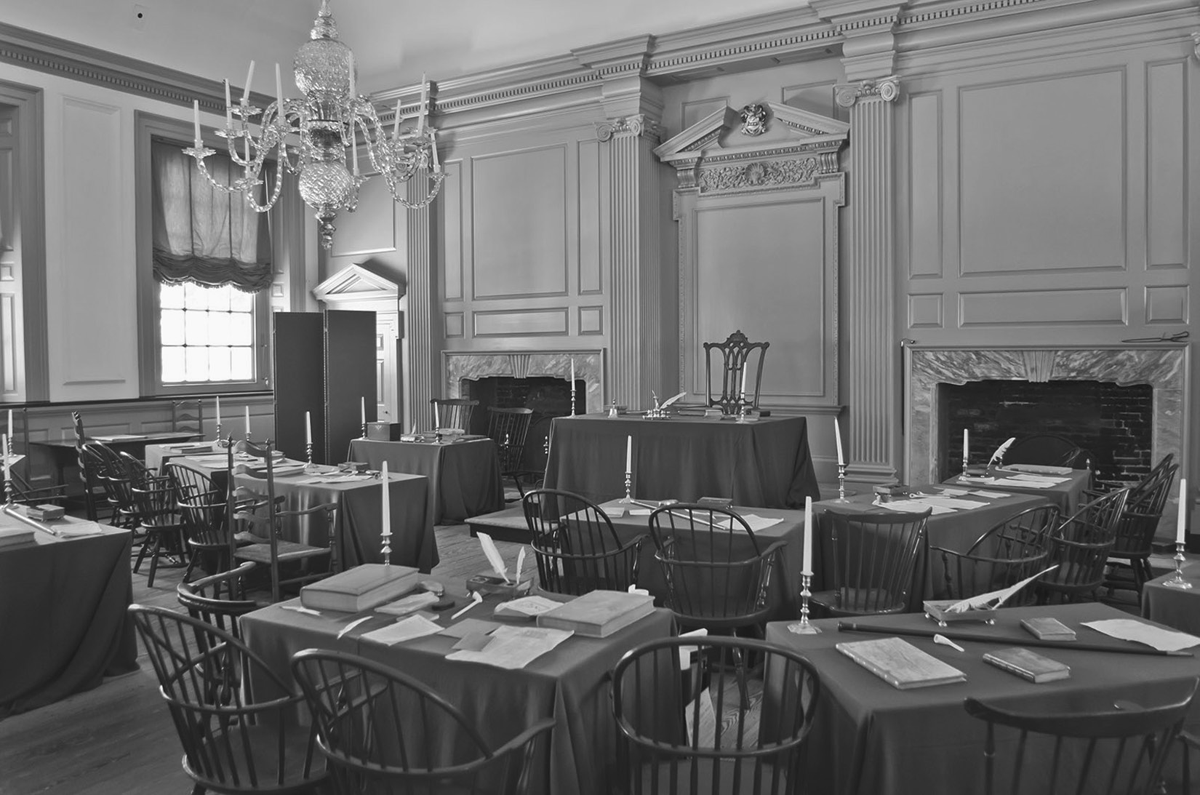 A contemporary photograph by Antoine Taveneaux of Independence Hall staged to look like it did when the delegates of the original 13 states met and drafted the US Constitution. There are several delegate tables covered in green tablecloths throughout the room. They are accompanied by wooden chairs and adorned with quill ink pens, candles, walking sticks, pipes, old books, and pieces of paper. The tables are all facing the table at the front of the room where George Washington would have sat. There are fireplaces on either side of Washingtonâ€™s desk.