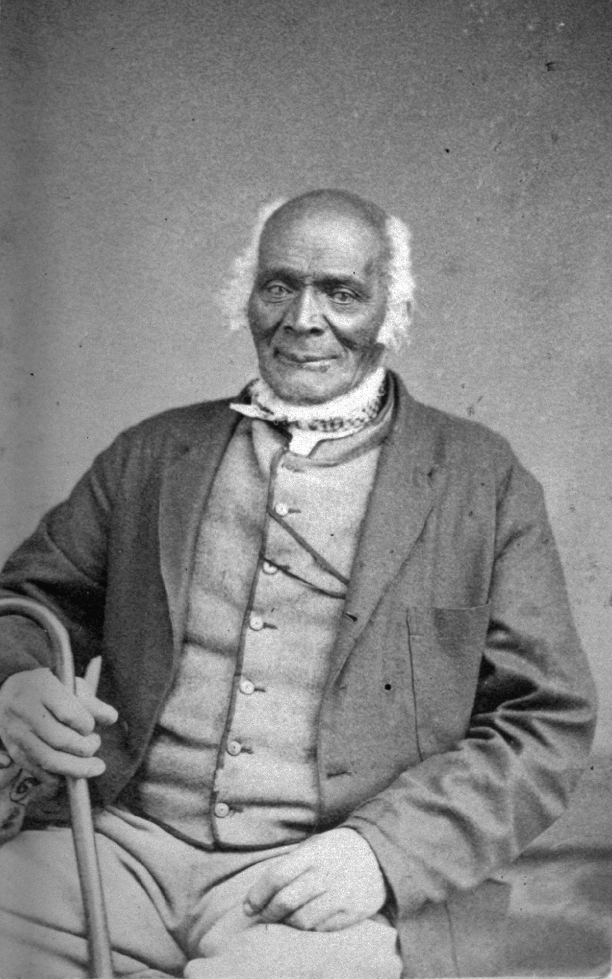 This photograph portrait of James Mars in 1870 depicts an elderly Black man with a fringe of white hair around a bald head, seated while holding a cane in his right hand. He wears a dark coat over a lighter vest and pants.