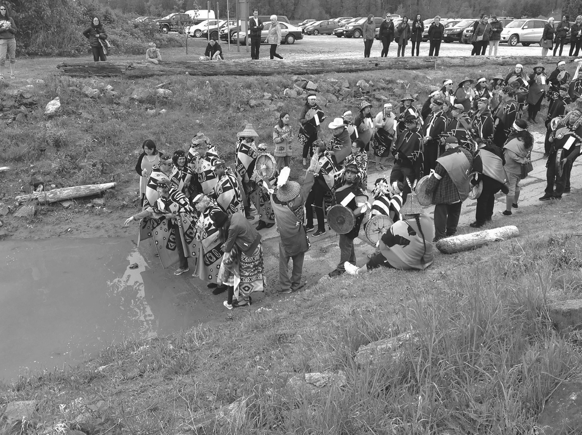 This photograph shows a Kwantlen First Nation salmon ceremony ritual, where the bones from the first salmon harvest are returned to the water by elders and community members dressed in brightly colored ceremonial clothing. Others nearby play drums.