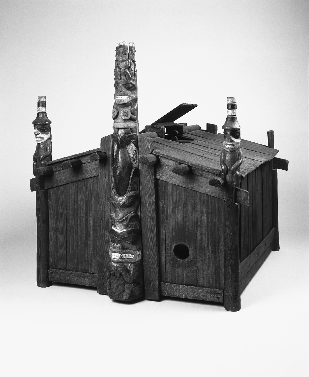This nineteenth-century model is a small reproduction of a Haida chieftainâ€™s house. It is made of a rich dark brown wood and has a totem pole rising out of the front of the model depicting a sea wolf, whale, a female shaman, and three watchmen. These houses were used in the winter for potlatch ceremonies.