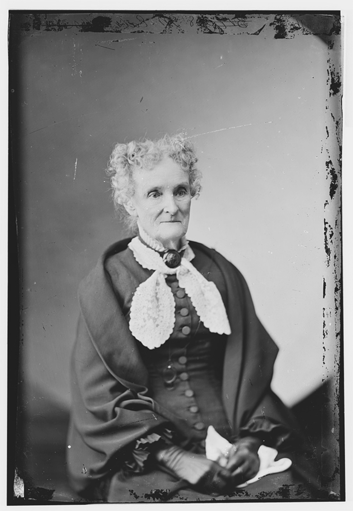A circa 1870â€“1880 black-and-white photograph of Margaret Eaton. She is an elderly woman with curly gray hair, wearing a dark dress with a white scarf and dark gloves. She is seated and turned slightly to her left, looking in that direction.