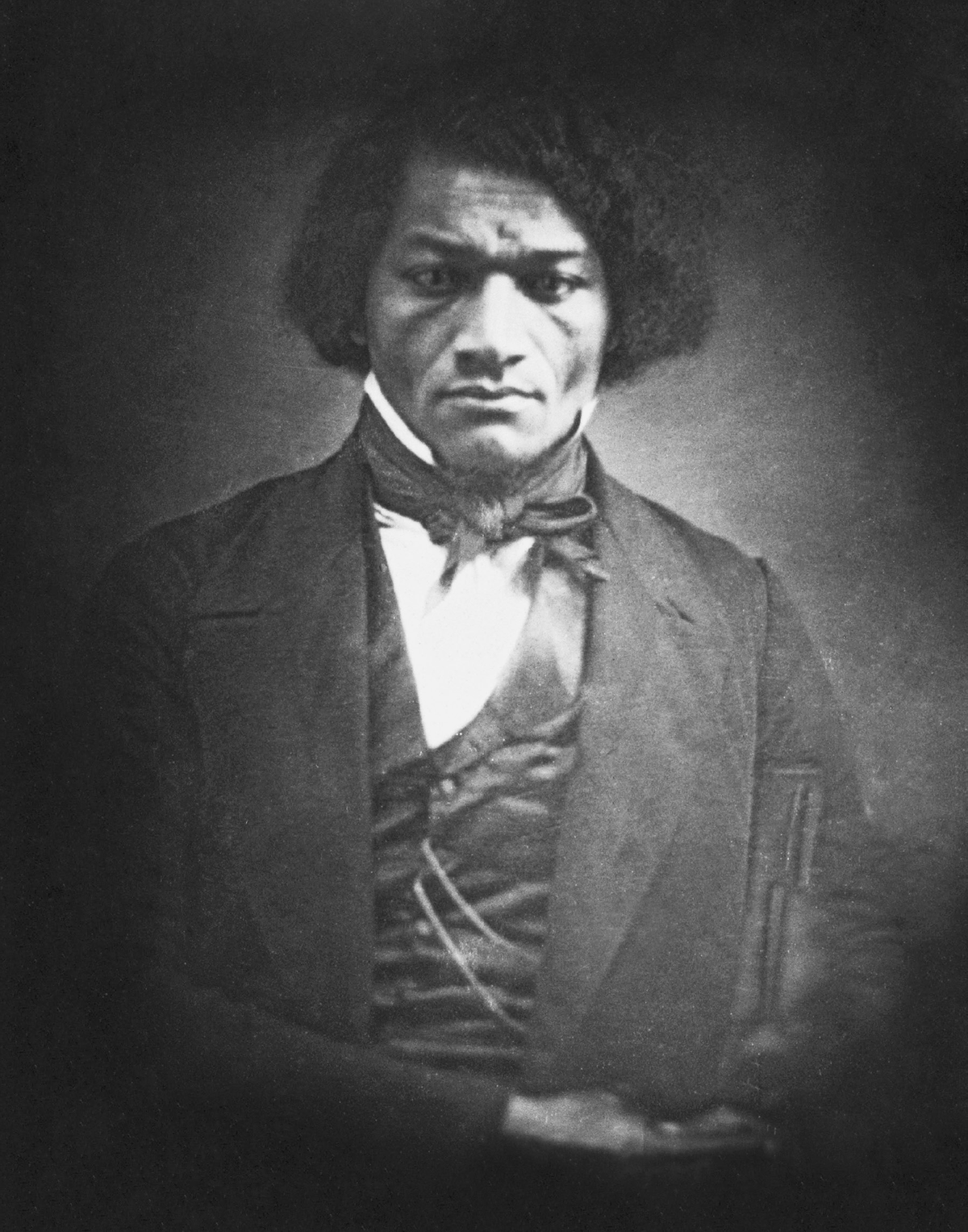 This is a photograph portrait of Frederick Douglass as a young man around 1847. A man with dark, tightly curled, medium-length hair wears a white shirt under a dark coat and vest, with a dark cloth tied at his neck. He faces and looks forward at the viewer.