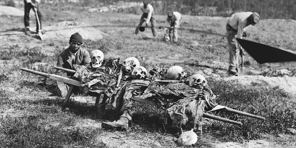 A stark photograph from 1865 shows a Black man crouched and solemnly posing next to a collection of skulls and bones. Other Black men are seen in the background with shovels and bending over to collect items from the ground.