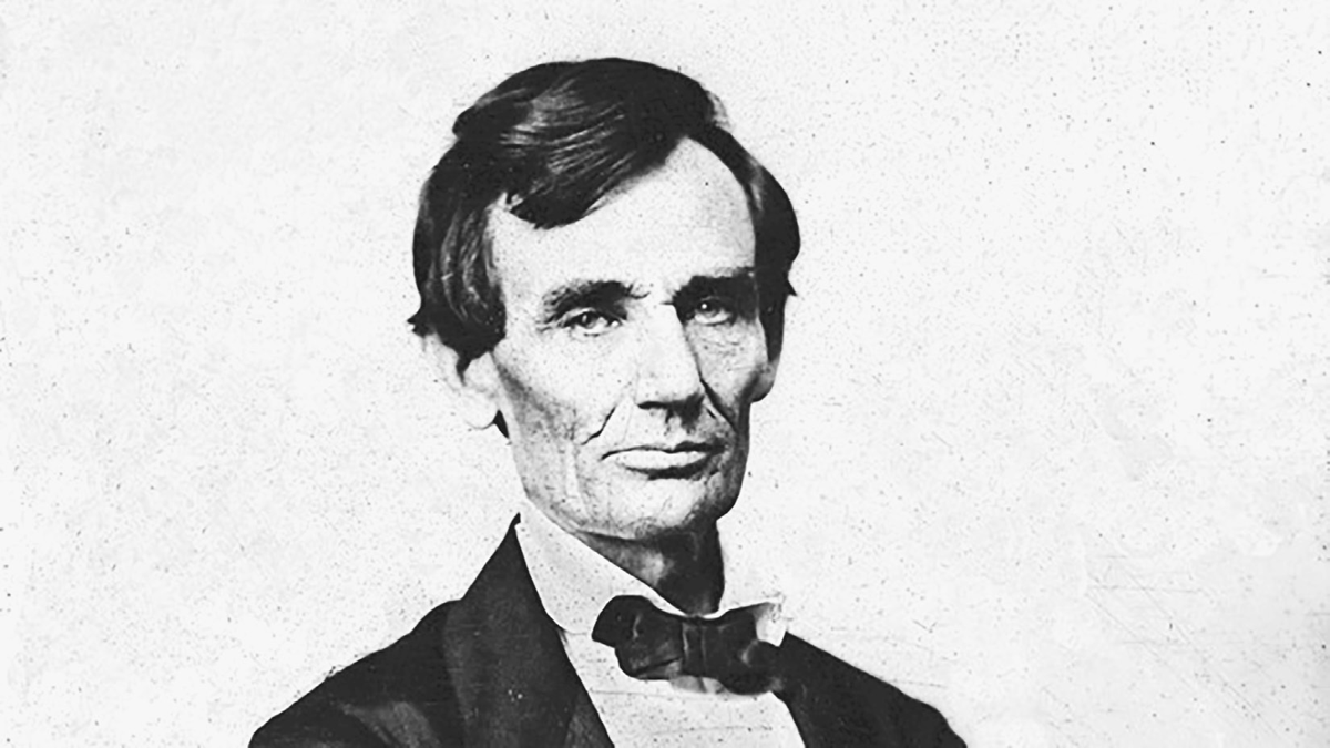 An 1860 photograph of Abraham Lincoln wearing a formal suit and black bow tie. He has dark hair hanging over a very high forehead, prominent cheekbones, and a very square jaw. He is turned slightly to his left but faces the viewer.