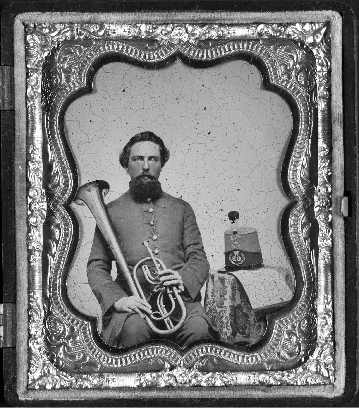 An unidentified soldier clad in gray holds an instrument similar to a French horn. The photograph is placed in a cover to a book that is ornately bound with red trim.