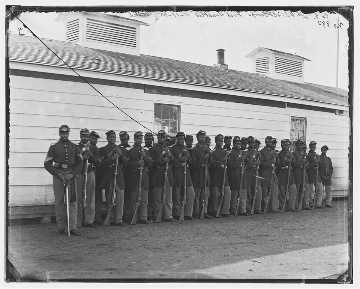 A photograph by William Morris Smith of Black men in Union uniforms standing in a row in front of a building that may be a barracks. They stand with guns held in front of them with the butts resting on the ground.