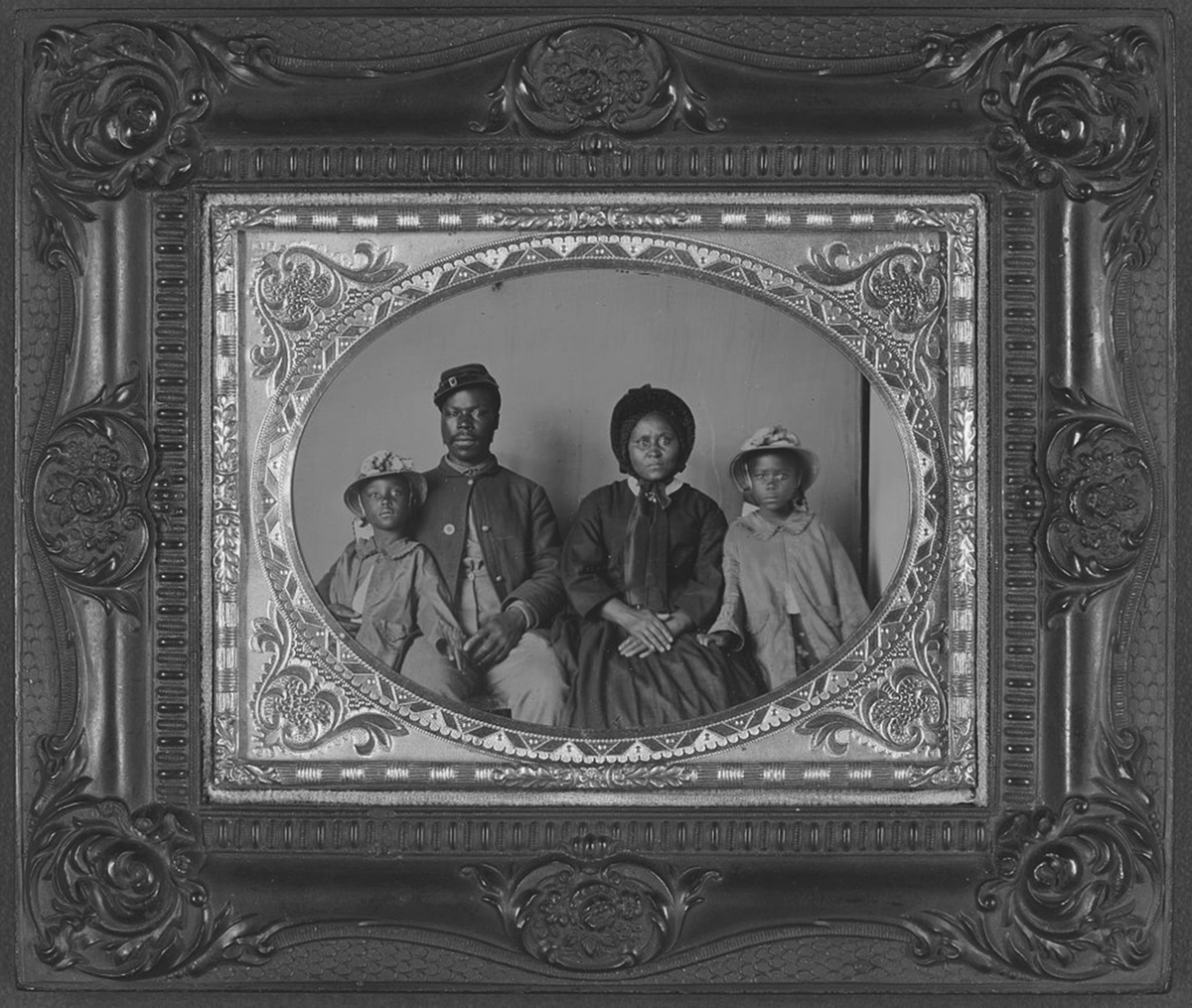 This African American family dressed in their finest clothes for this photograph, projecting respectability and dignity that was at odds with the Southern perception of Black Americans. The father wears a US Colored Troops uniform, while the mother and two daughters are dressed in dresses and hats. The parents are seated with the children standing on either side of them.