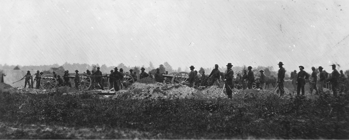 A sepia-toned 1864 photograph of Union soldiers from the Pennsylvania Light Artillery with their cannons at Petersburg, Virginia. Dozens of men stand in the middle distance with what appears to be trees in the distance.