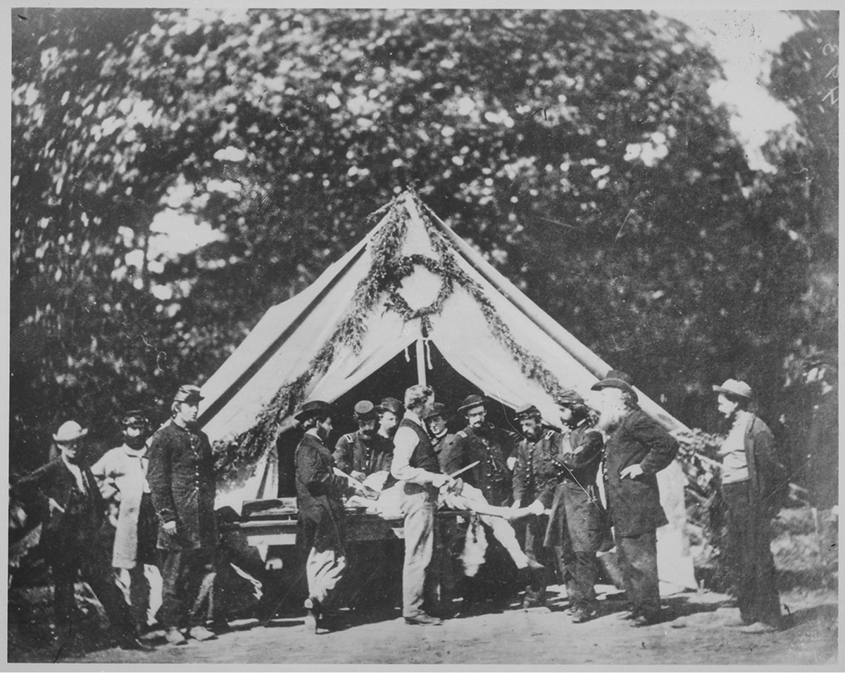 An 1863 photograph at Gettysburg of a medical tent with about a dozen men huddled around a body on a table. A man in the foreground holds what appears to be a saw or knife.