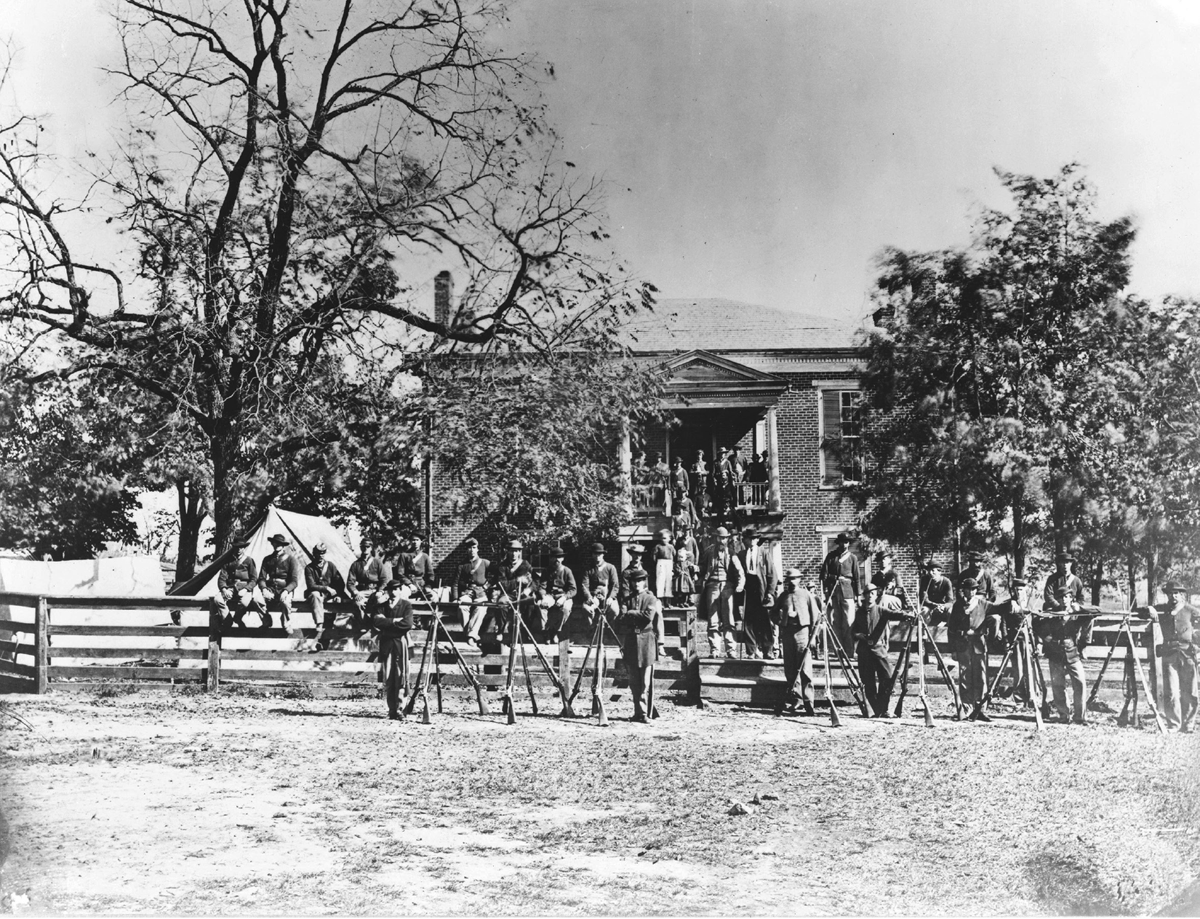 Union soldiers pose in front of the Appomattox Court House after Leeâ€™s surrender in April 1865. The men stand in front of their weapons, which are displayed together. The courthouse looms in the background.