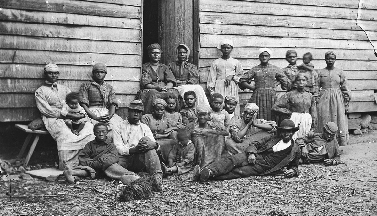 An 1862 photograph of over a dozen African American â€œContrabandsâ€ sitting or standing in front of a cabin in Virginia. They range in age from children to the elderly. The women wear long dresses and head scarves. The men wear shirts, trousers, and hats, and one also has a coat and vest. At least some of the children are barefoot.