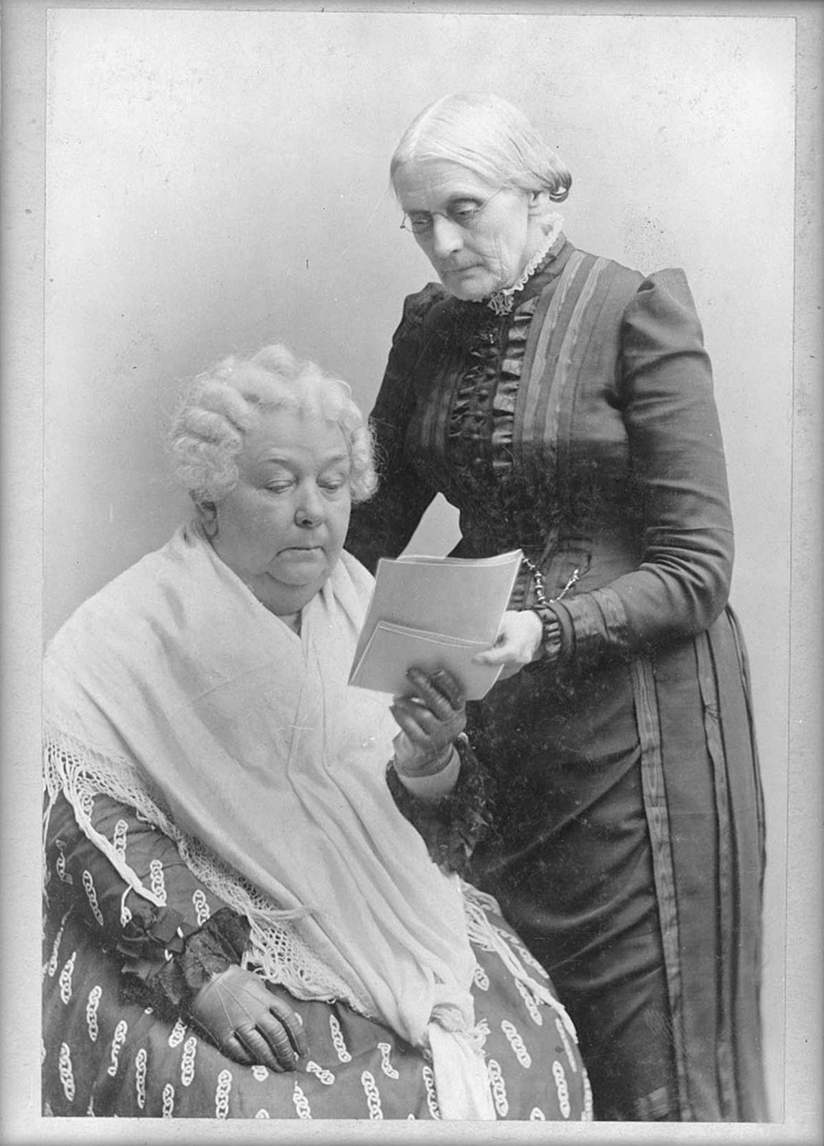 Photograph from between 1880 and 1902 of Susan B. Anthony and Elizabeth Cady Stanton. Both have white hair. Anthony wears glasses and a dark dress with some ruffles and inset stripes down the front, while Stanton has a dress with a large pattern and a white fringed shawl over her shoulders and torso. Anthony is standing at right, holding a document for Stanton, seated, to read.