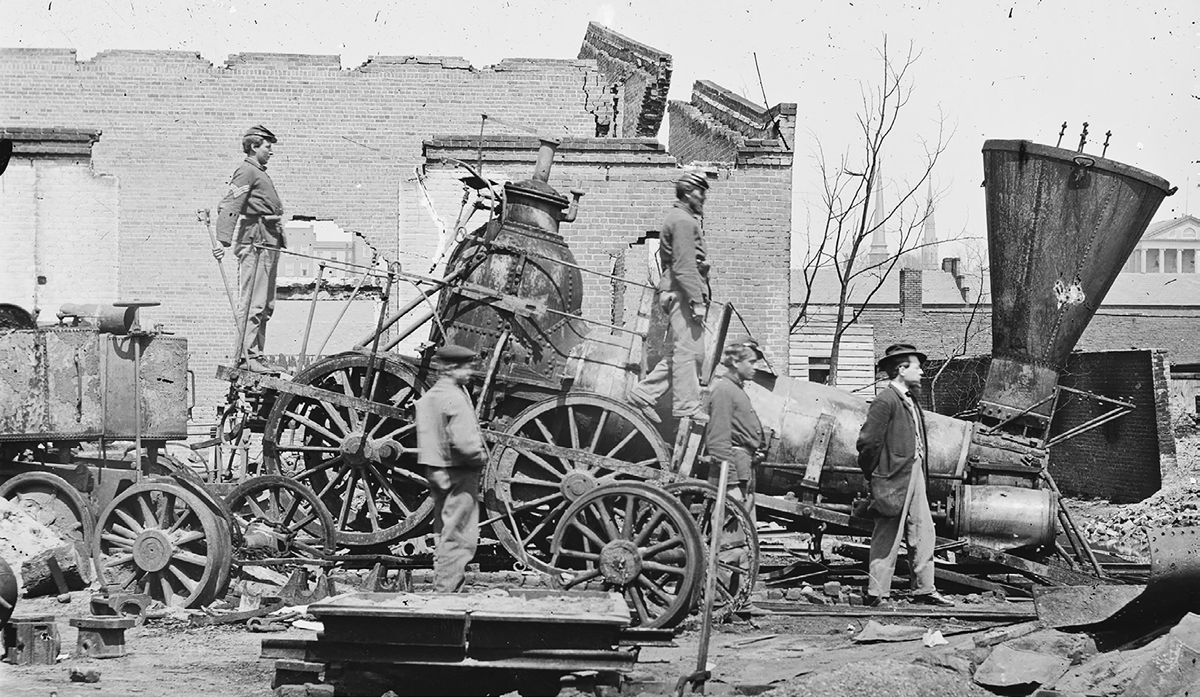 An 1865 photograph of a destroyed locomotive in Richmond, Virginia. Four men stand on and near it amid other rubble. A destroyed building is in the background.