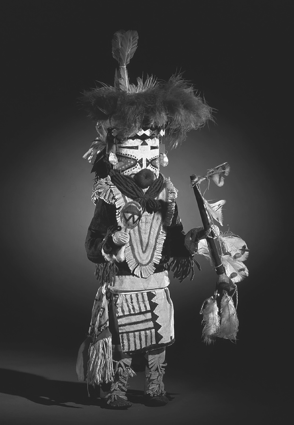 This Zuni kachina doll is made of brown feathers, and wears knee-high moccasins in a light cream color and painted clothing. He sports a bright red round mouth. He holds a rattle in one hand and another object, possibly a pipe adorned with feathers in the other.