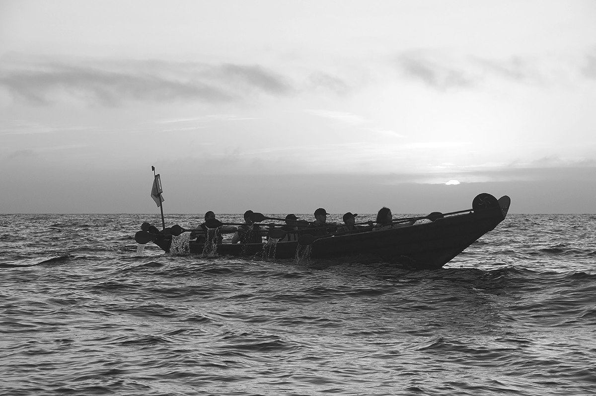 A beautiful sunrise in shades of orange serves as the backdrop of a photograph of Chumash people crossing from the Channel Islands Harbor to Santa Cruz Island off the coast of California in a traditionally built tomol, a plank canoe. The boat contains rowers.