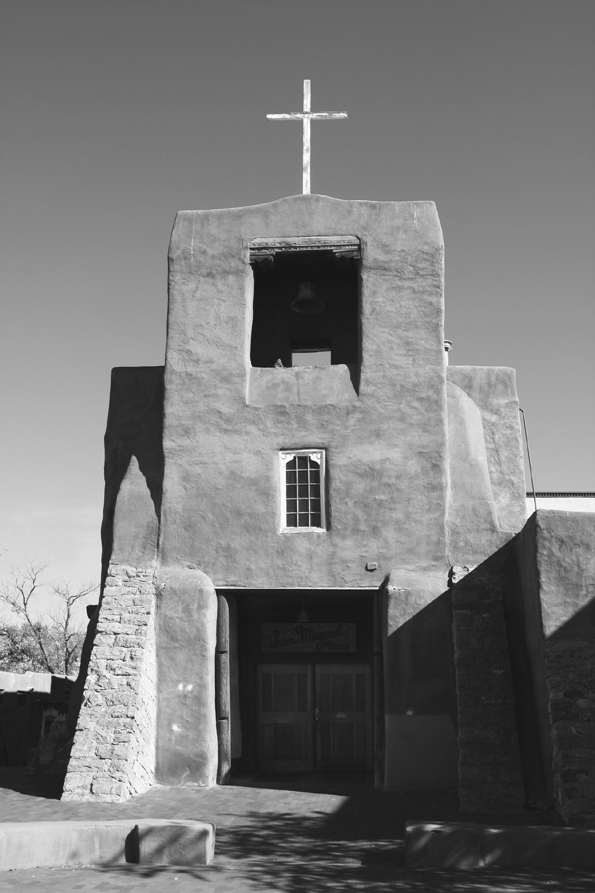 A photograph of the San Miguel Chapel in Santa Fe, New Mexico, built in 1610, the oldest Catholic church in the United States. Built of adobe, its rectangular architecture was typical of Franciscan frontier churches. The chapel includes a bell tower and cross on top. Small stones adorn the front sides of the building and a small window in the top to allow light to enter.