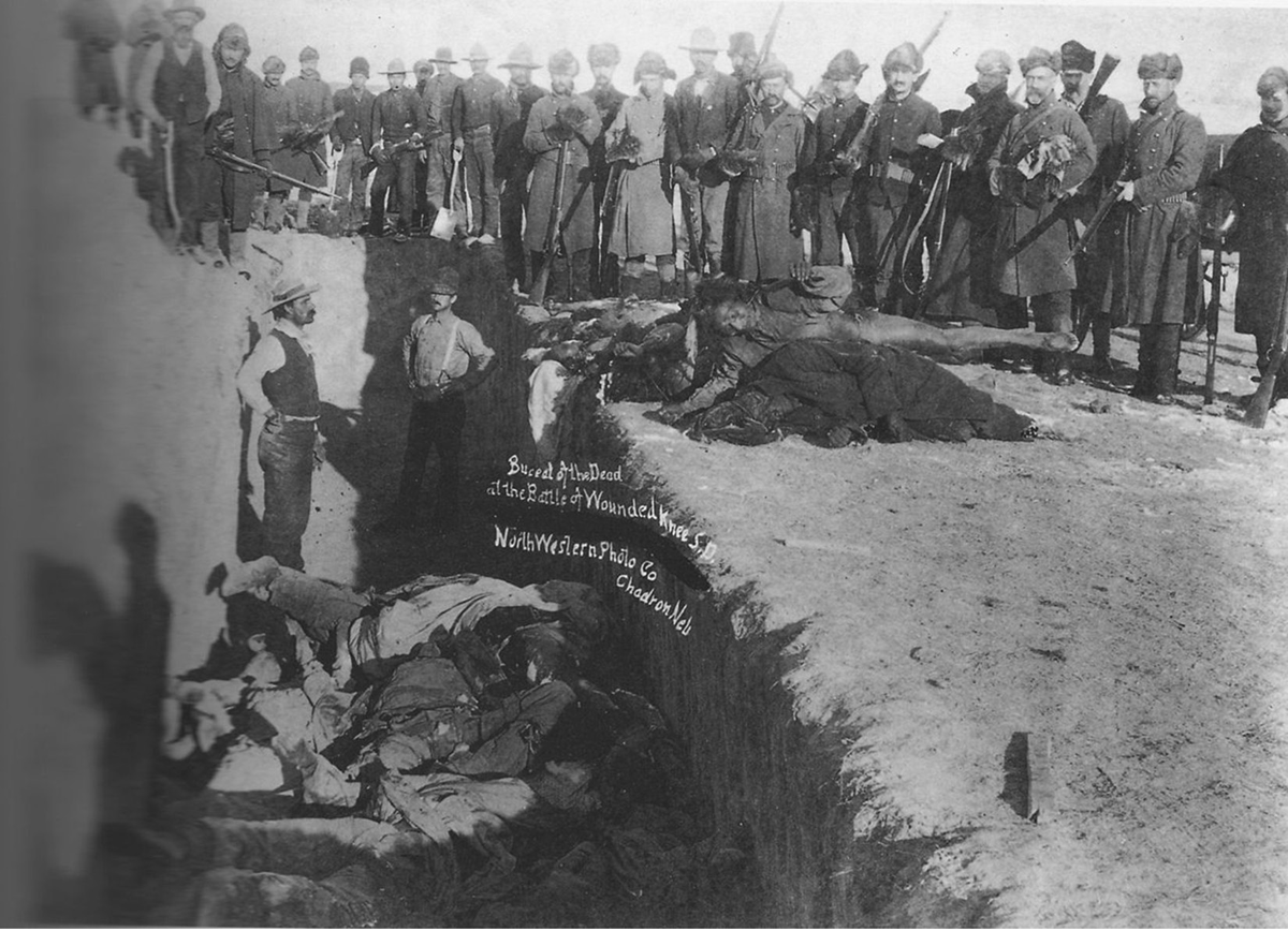Soldiers stand around a mass grave with piles of bodies in piles in and out of the grave. On the image is written, â€œBureal of the Dead at the Battle of Wounded Knee S.D. NorthWestern Photo Co., Chadron, Neb.â€