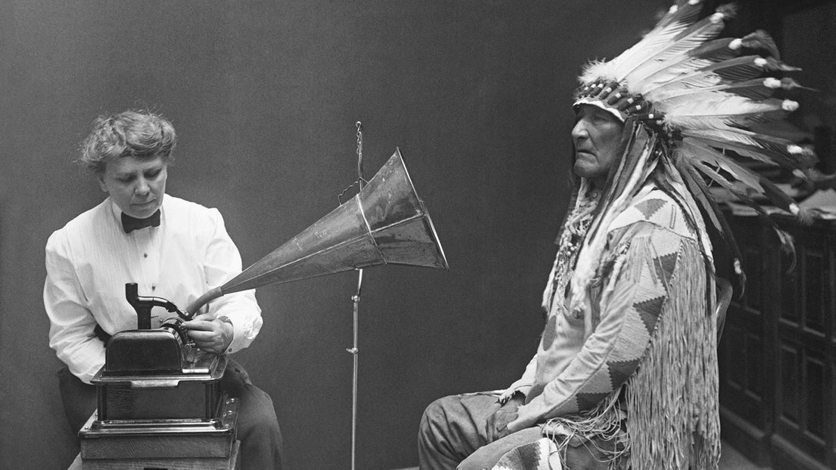 An old man in traditional Indigenous clothing stands before an audio device tended by a woman in a shirtwaist.