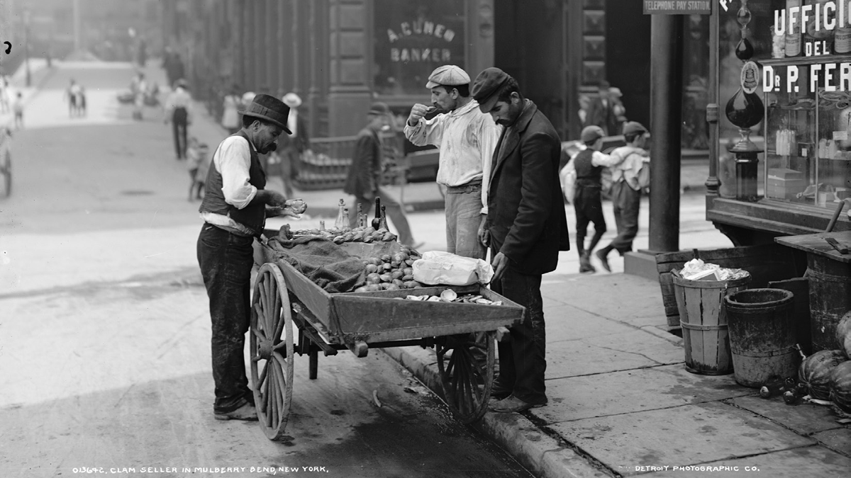 A man sells clams from a cart in an urban setting.