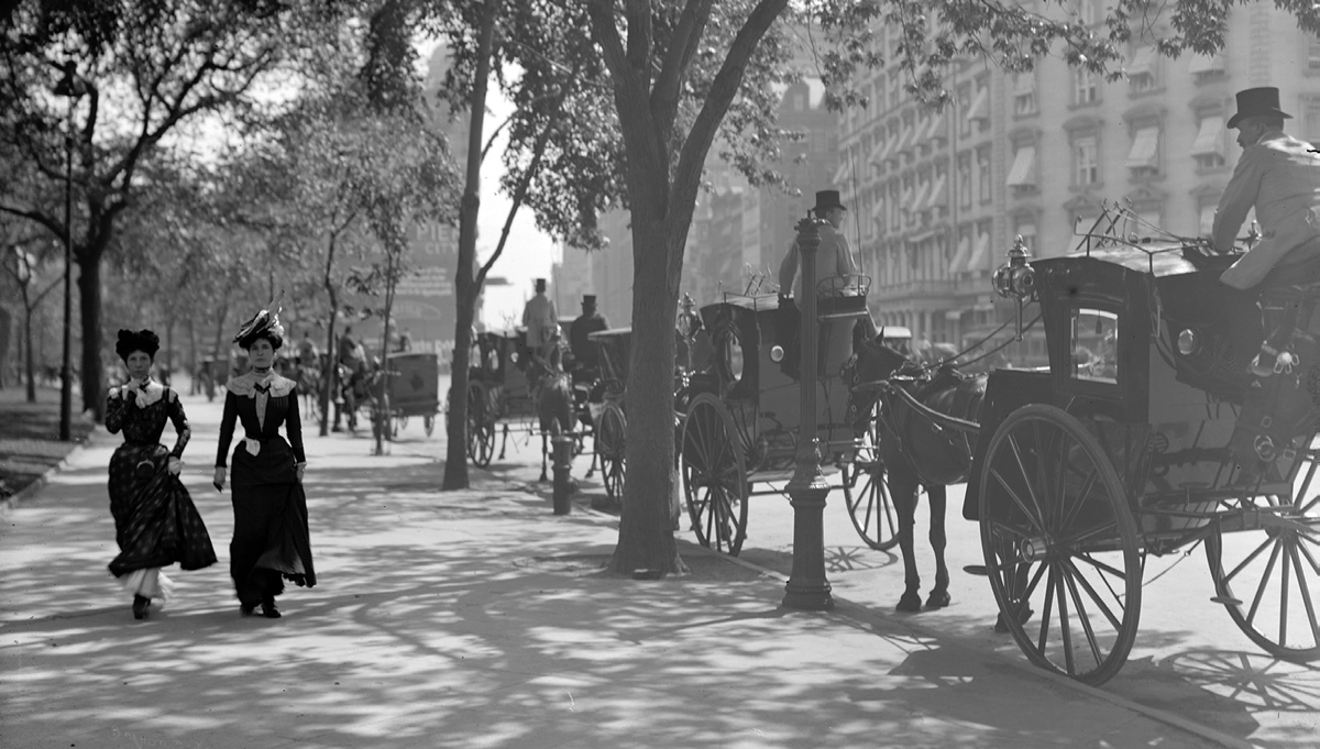 Two women pass by a line of horse-drawn carriages.