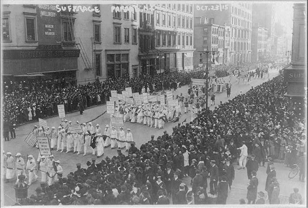 Women dressed in white march in a parade with throngs of people crowded on both sides of the street.