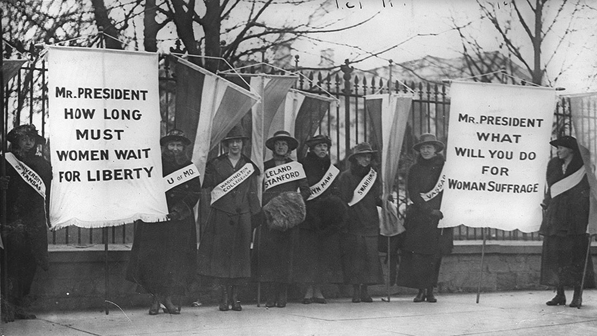 Women wear sashes indicating their alma maters as they picket the White House. Two large banners near them read â€œMr. President how long must women wait for libertyâ€ and â€œMr. President what will you do for woman suffrage.â€