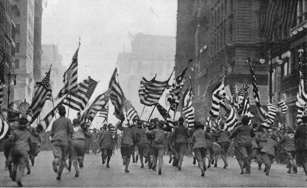Young boys in boy scout uniforms run down a city street carrying American flags.