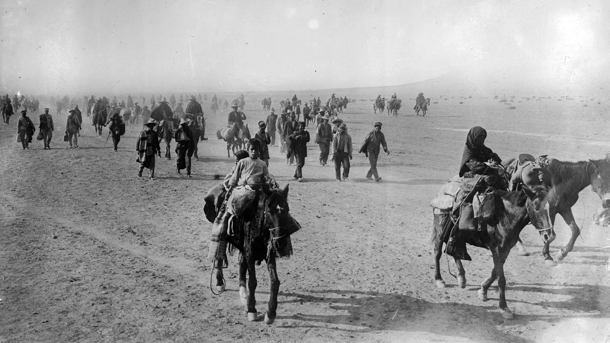 Refugees, some riding what appear to mules, but most walking, move through a desert.