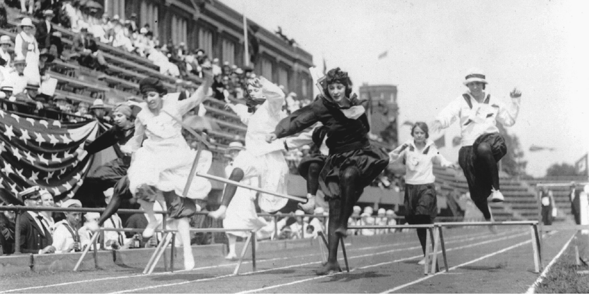 Women in dresses jump over hurdles on a track.