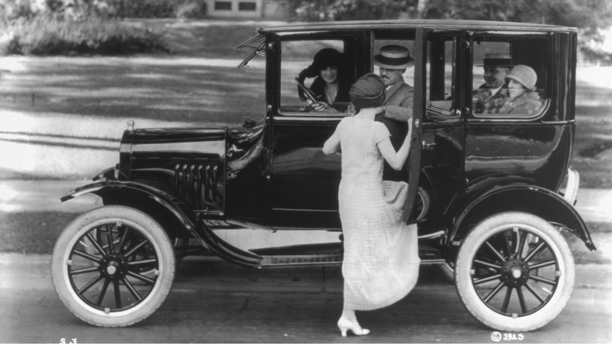 A woman stands in front of an open car carrying two men and two women.