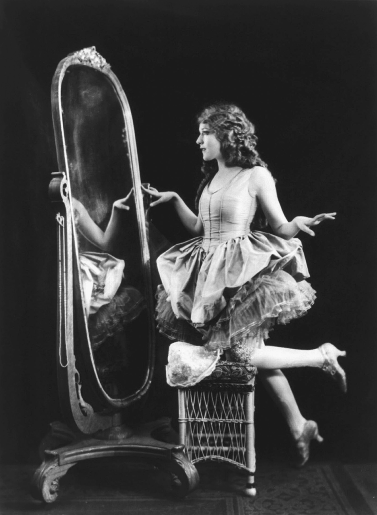 A woman in makeup and wearing a dress stands in front of a mirror.