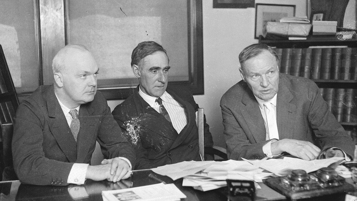 Three men sit at a desk.