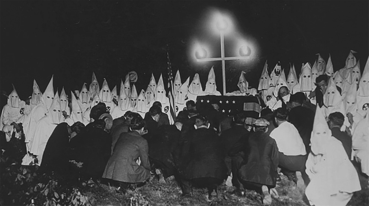 Men in Ku Klux Klan regalia pose for photographers under a cross.