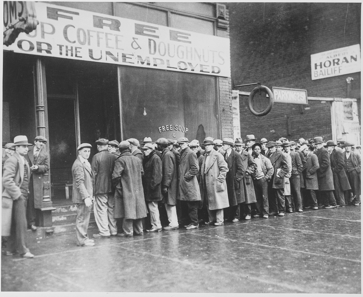 Men wait in line outside of a building advertising free coffee <?>and doughnuts for the unemployed.