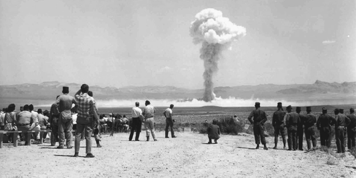 A crowd of soldiers and civilians watch a mushroom cloud rising in the distance.