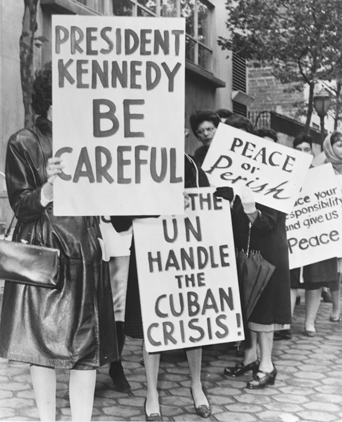 A crowd carrying signs reading â€œBe Carefulâ€ line a city street.
