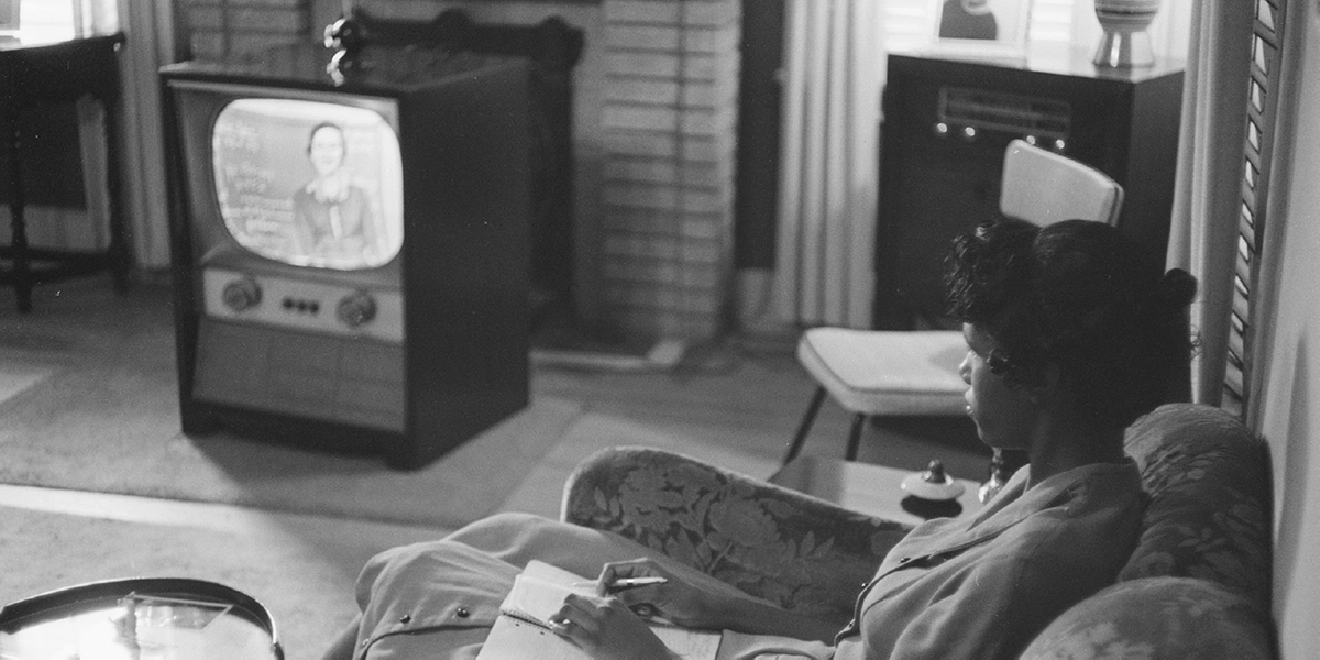 A young woman sits watching a woman in front of a chalkboard on television.