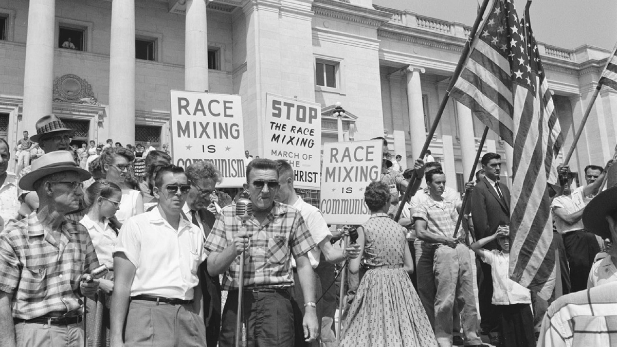 Protestors outside of a government building hold American flags and placards reading â€œRace Mixing Is Communism.â€