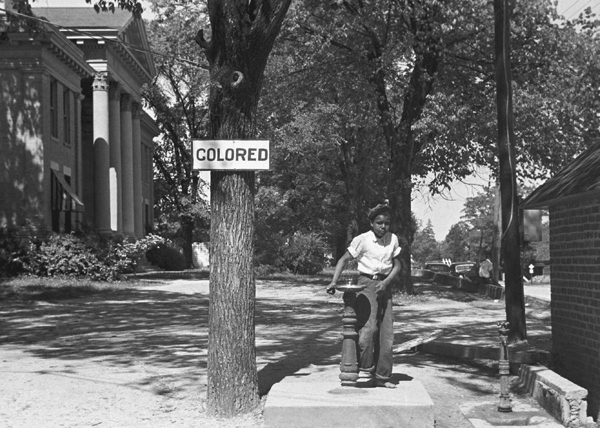 A boy drinks from an outdoor water fountain in front of a tree with a sign reading â€œColored.â€