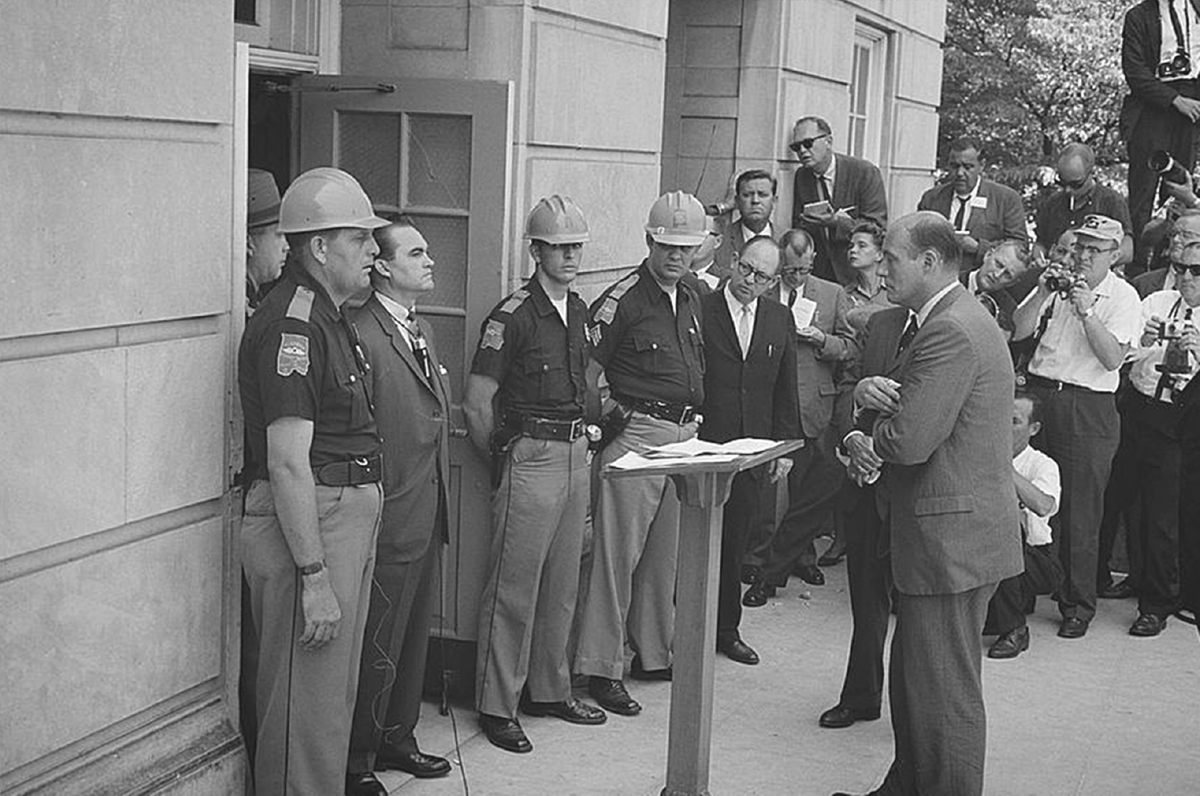 A man stands in front of a door, flanked by law enforcement officers, as another man stands before him.