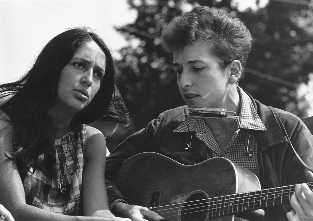 A young woman and a young man sit, holding a guitar and harmonica, respectively.
