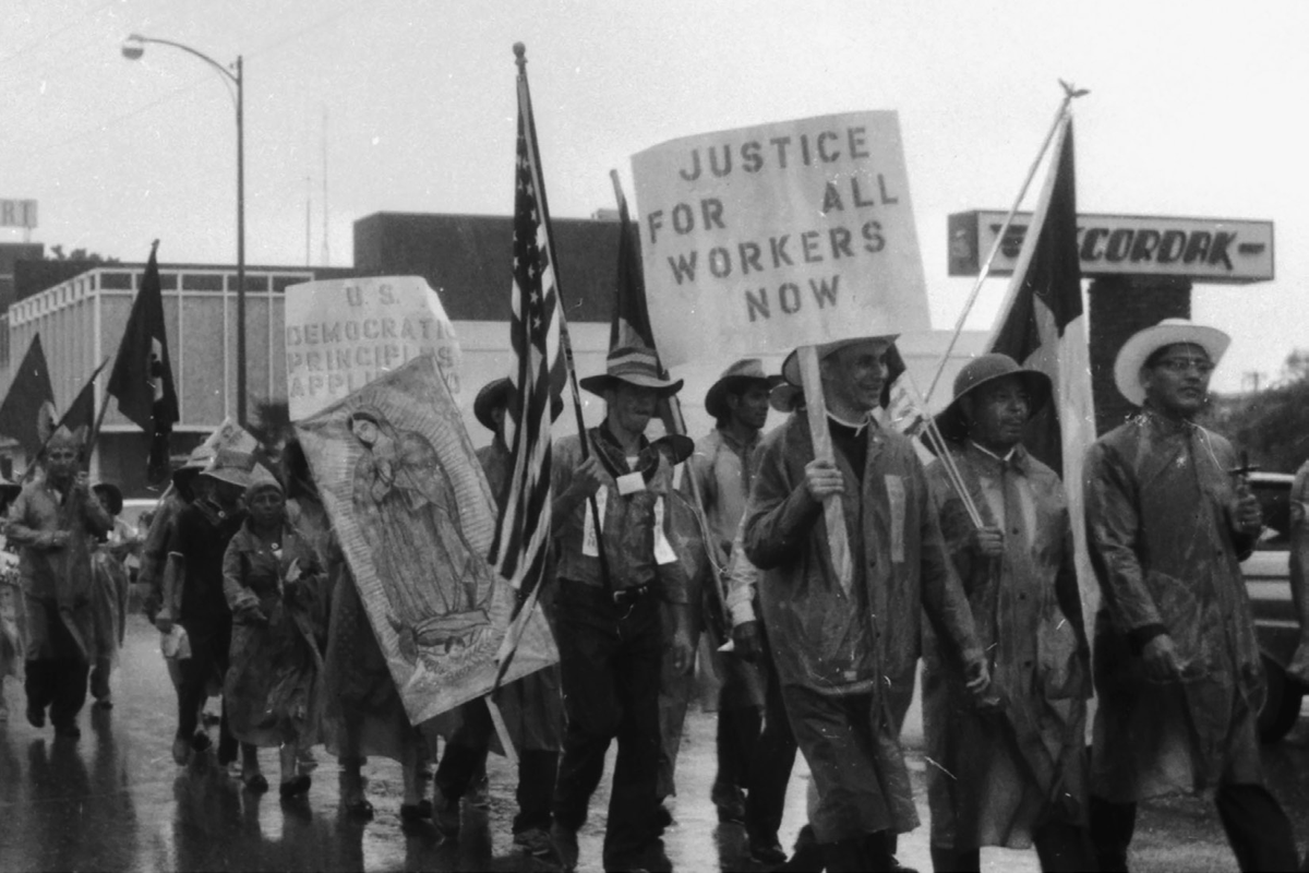 Menâ€”including at least one Catholic clergymanâ€”and women march through a wet street, carrying a sign reading â€œJustice for all workers nowâ€ and an image of the Virgin Mary.