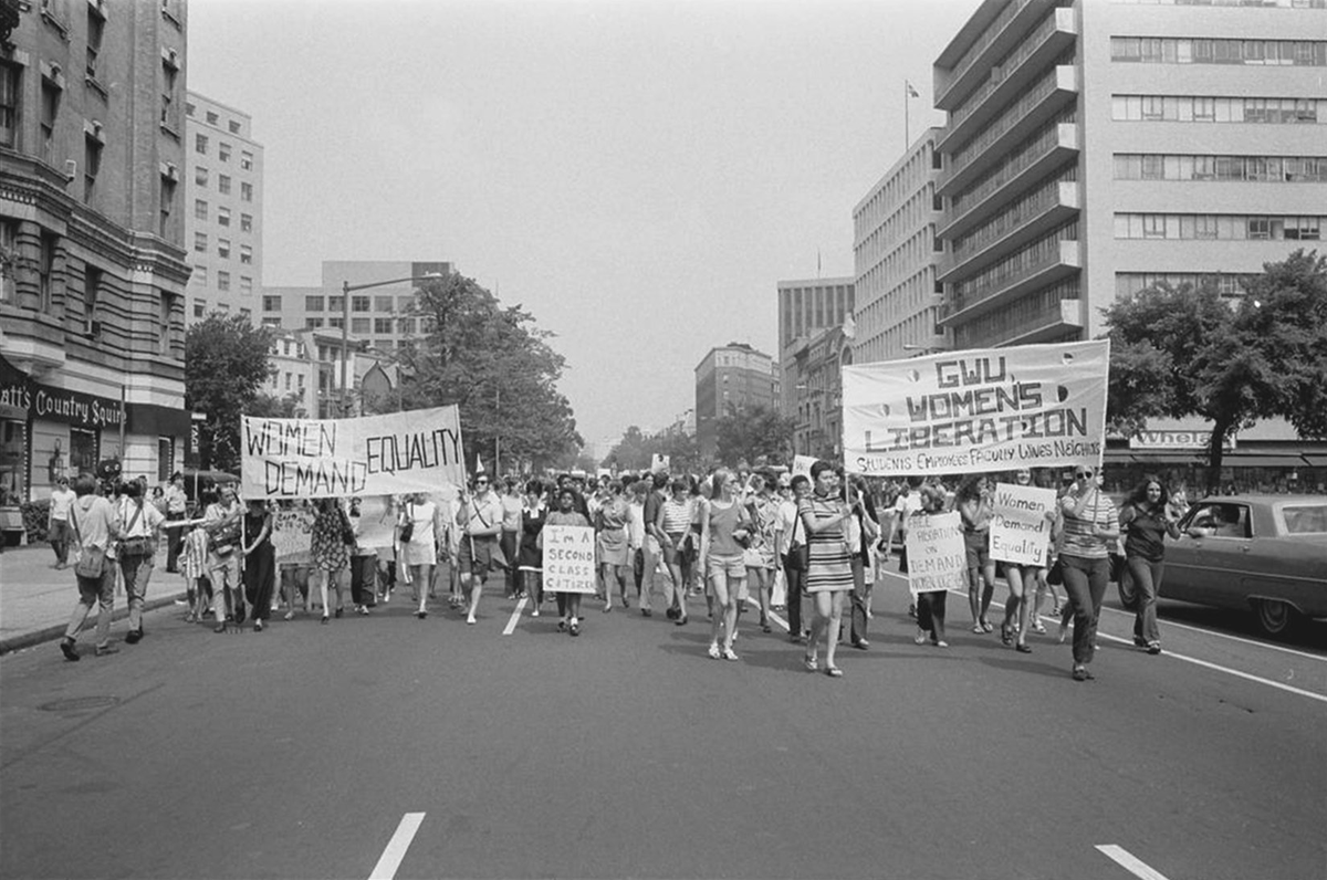 Women march down a city street with large banners reading â€œWomen Demand Equalityâ€ and â€œGWU Womenâ€™s Liberation.â€