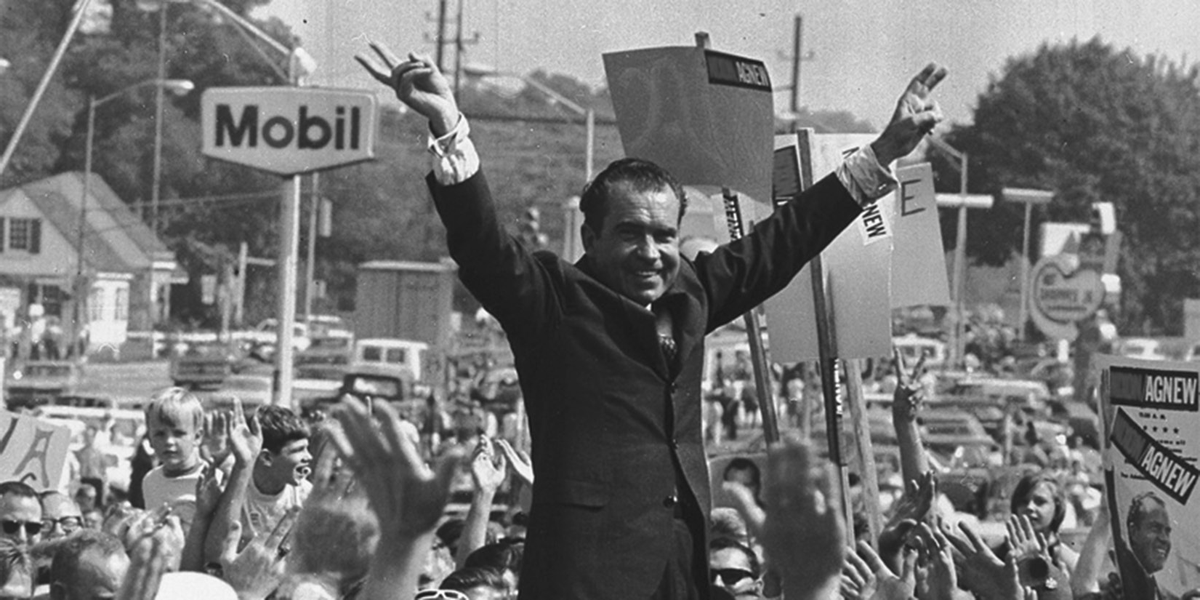 A man smiles and raises both arms overhead amid a cheering crowd.