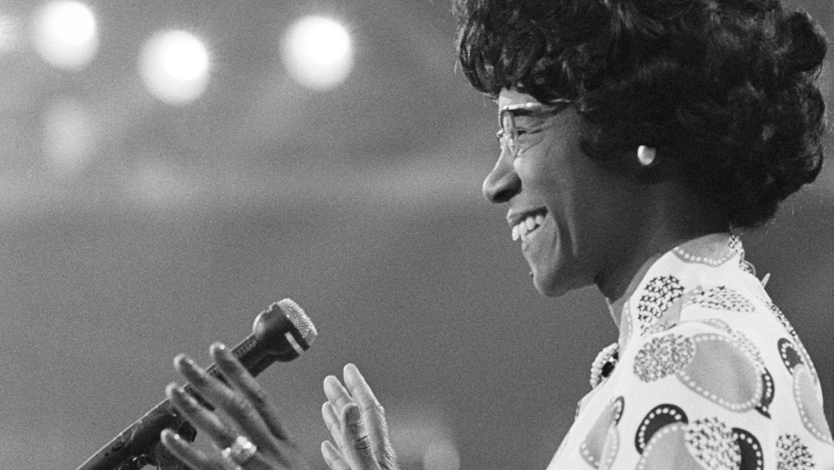 A woman, depicting in a side profile, smiles before a microphone.