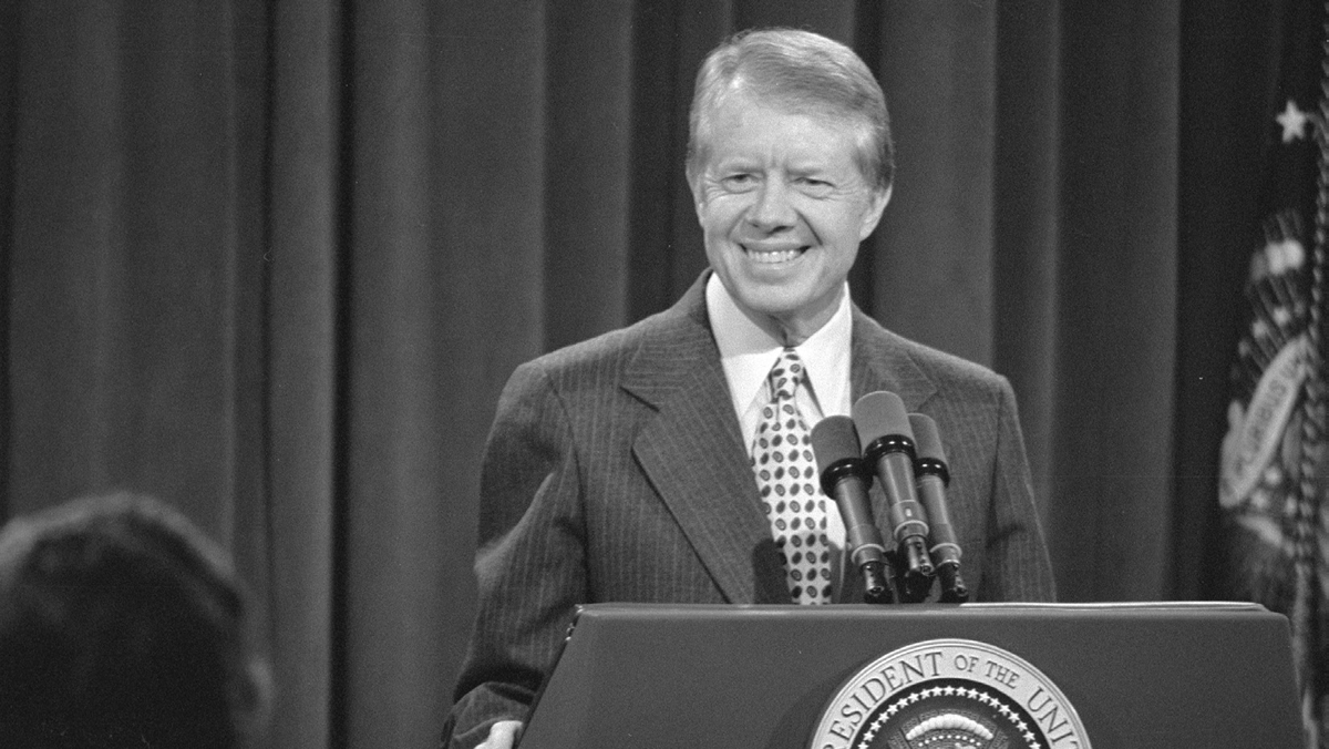 A man in a suit stands before a lectern with microphones.