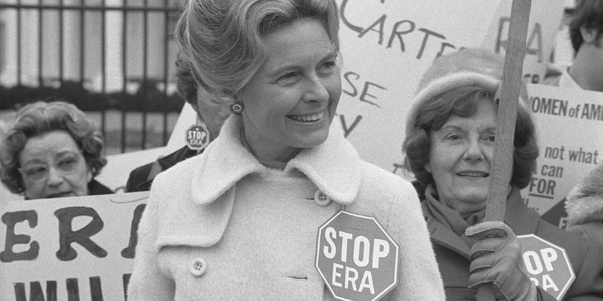 A woman speaks before a microphone as demonstrators hold signs reading â€œStop ERAâ€ behind her.